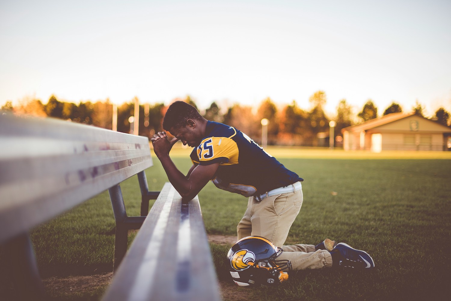 Football player neiling in front of a bench saying a prayer.